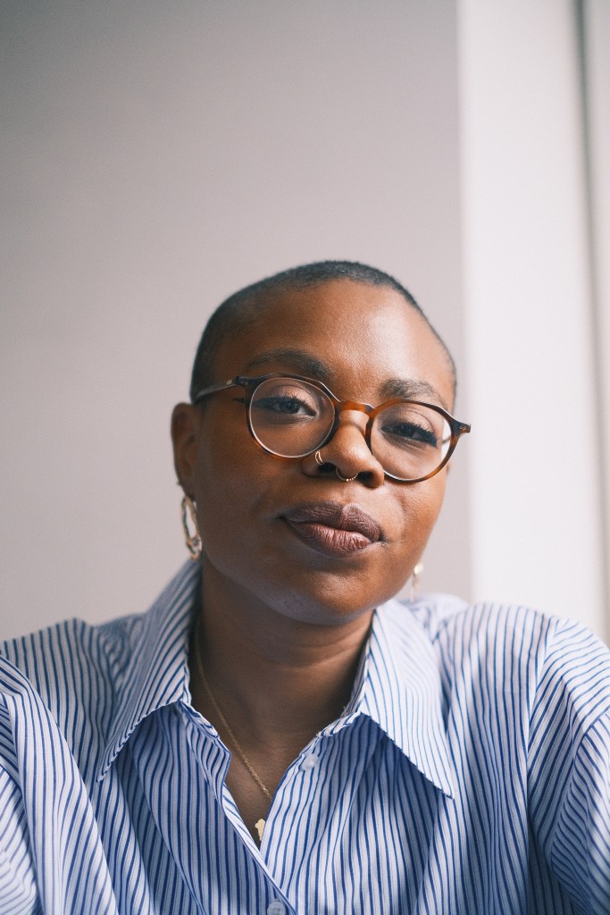 A picture of a Black woman with a shaved head smiling at the camera with her mouth closed, she is wearing glasses, gold earrings and a white and blue stripped shirt dress